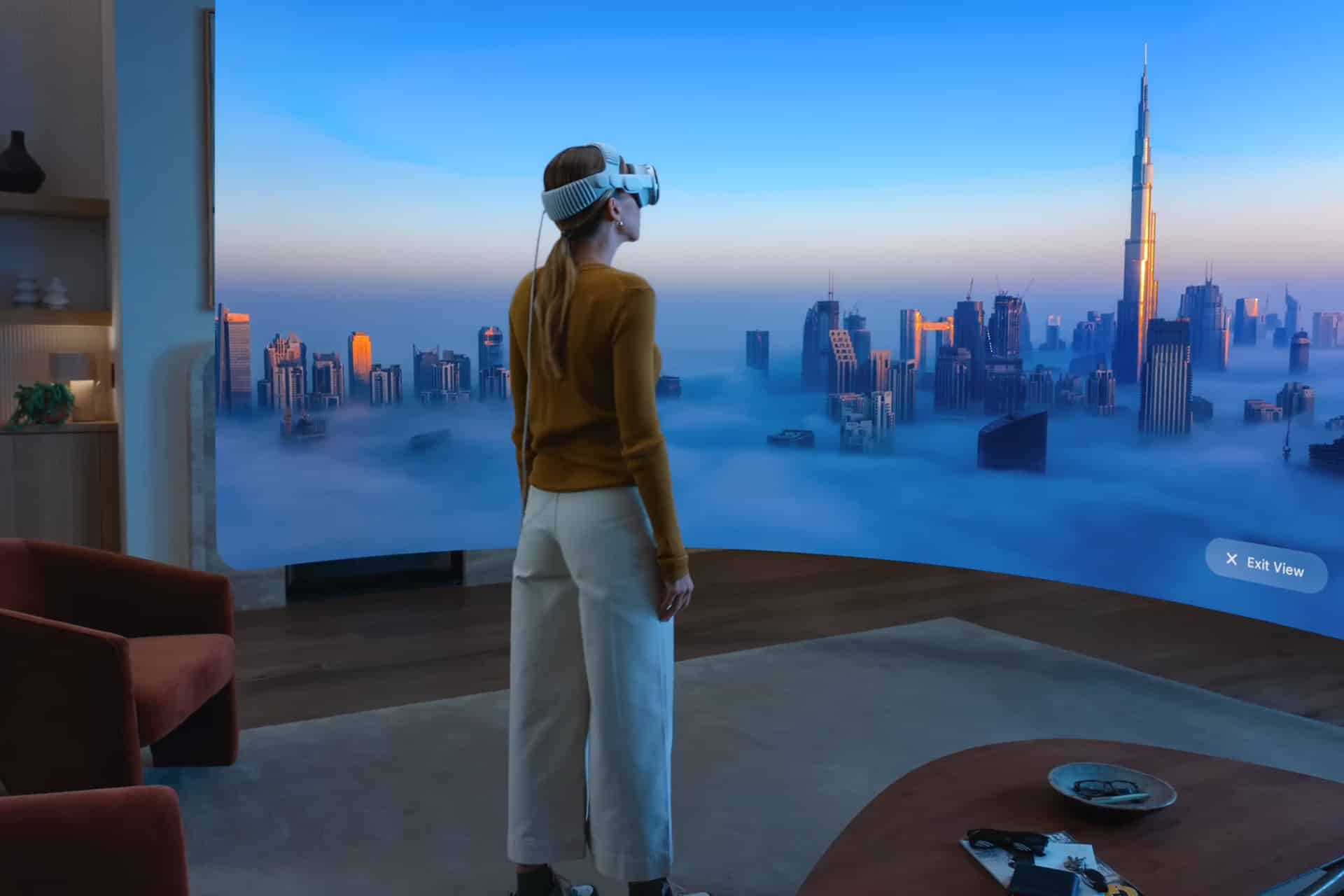 A woman wearing a VR headset stands in a modern living room, viewing an immersive cityscape in the Spatial Computing Era, with tall skyscrapers and clouds on a large curved screen. The tallest building resembles the Burj Khalifa.