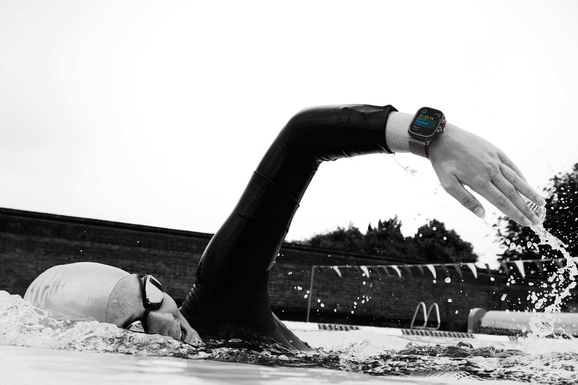 Apple Watch Water Lock - A swimmer wearing a swim cap, goggles, and an Apple Watch Scuba swims freestyle in an outdoor pool, with one arm outstretched above the water. The smartwatch display is clearly visible.