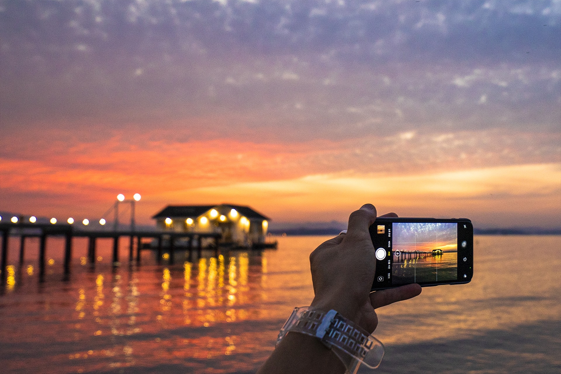A hand holds a smartphone, seamlessly integrated into the Apple ecosystem, capturing a photo of a pier and illuminated building over calm water at sunset, with vibrant orange and purple hues reflected softly on the surface.