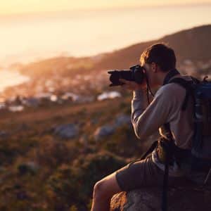 A person wearing a backpack sits on a rock at sunset, taking photos of a scenic, blurred landscape with hills and water in the background—capturing every moment seamlessly within the Apple ecosystem.