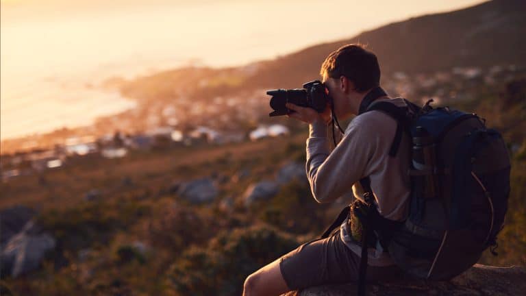 A person wearing a backpack sits on a rock at sunset, taking photos of a scenic, blurred landscape with hills and water in the background—capturing every moment seamlessly within the Apple ecosystem.
