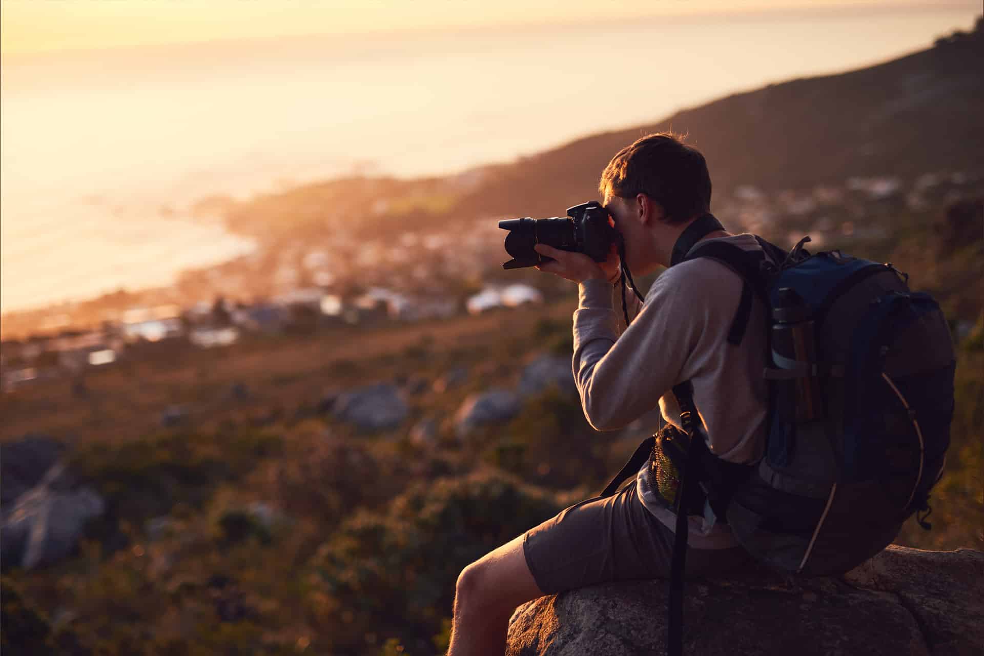 A person wearing a backpack sits on a rock at sunset, taking photos of a scenic, blurred landscape with hills and water in the background—capturing every moment seamlessly within the Apple ecosystem.