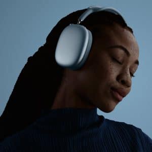 A woman with braided hair wearing large gray headphones and a pleated blue top has her eyes closed, appearing to enjoy the music, against a plain blue background.