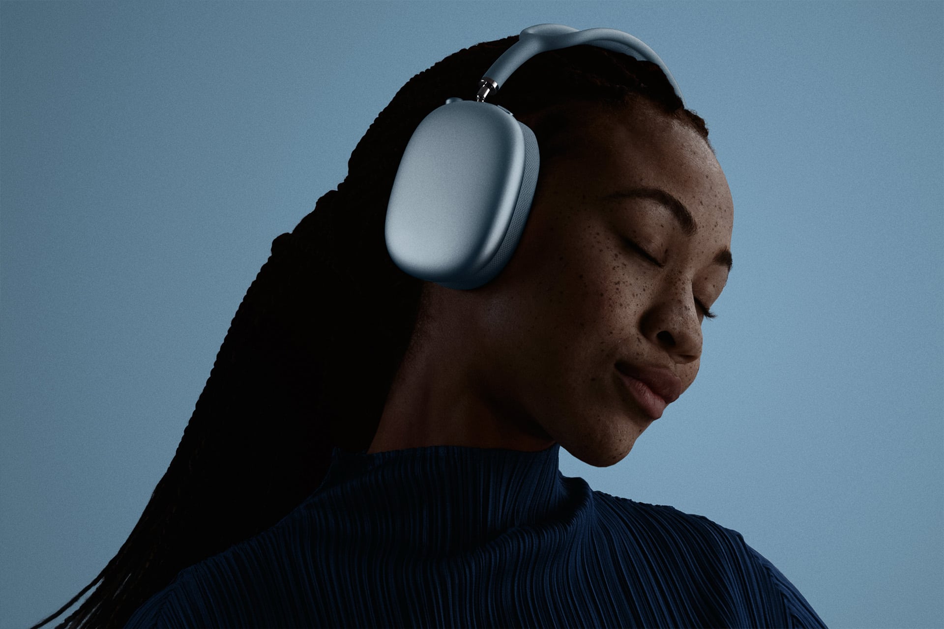 AirPods Max 2 - A woman with braided hair wearing large gray headphones and a pleated blue top has her eyes closed, appearing to enjoy the music, against a plain blue background.