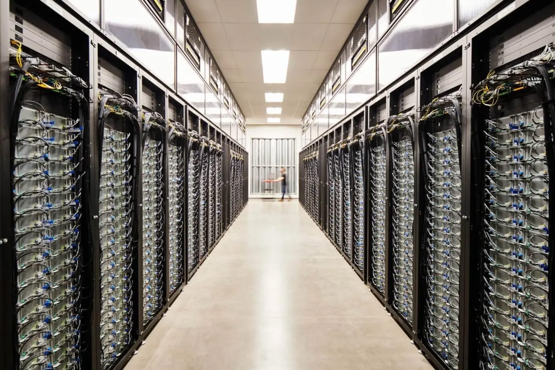 A brightly lit Apple data center hallway with rows of black server racks on both sides, filled with network equipment and cables, leading to a glass door at the far end.