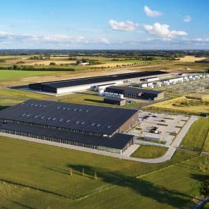 Aerial view of a large modern Apple data center complex surrounded by green fields and farmland, with multiple buildings, parking lots, roads, and electrical infrastructure under a blue sky with scattered clouds.