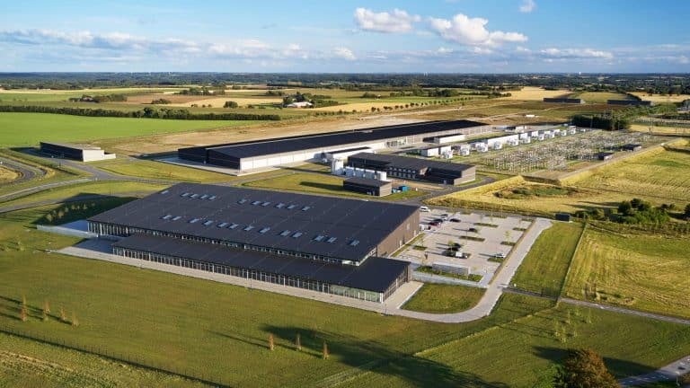 Aerial view of a large modern Apple data center complex surrounded by green fields and farmland, with multiple buildings, parking lots, roads, and electrical infrastructure under a blue sky with scattered clouds.