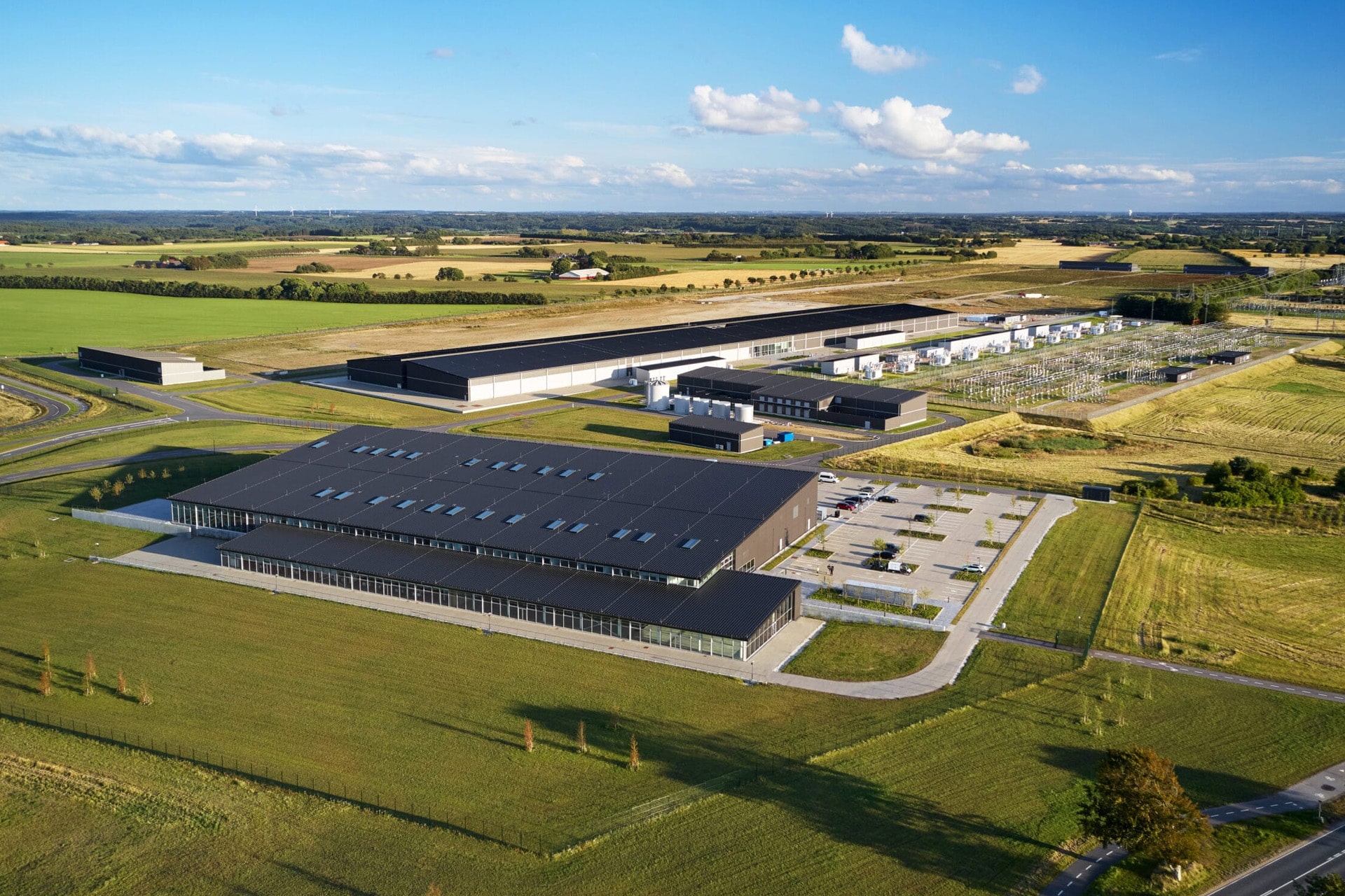 Aerial view of a large modern Apple data center complex surrounded by green fields and farmland, with multiple buildings, parking lots, roads, and electrical infrastructure under a blue sky with scattered clouds.