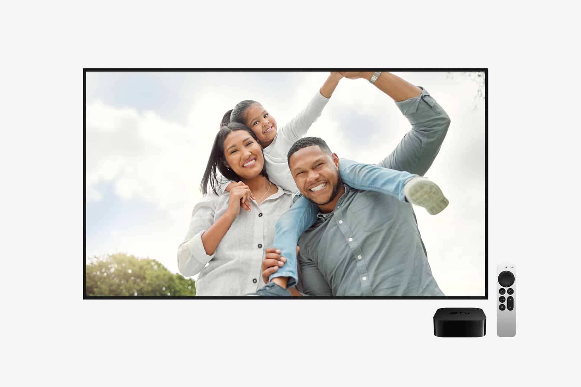 A happy family of three smiles while posing outdoors, displayed on a large TV screen reminiscent of Apple TV screensavers. In front of the TV are a remote control and a streaming device.