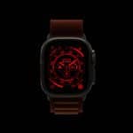 A smart watch with a dark case and brown band displays a red digital interface featuring various circular dials and numbers, all set against a black background—reminiscent of the Apple Watch Night Mode for low-light visibility.