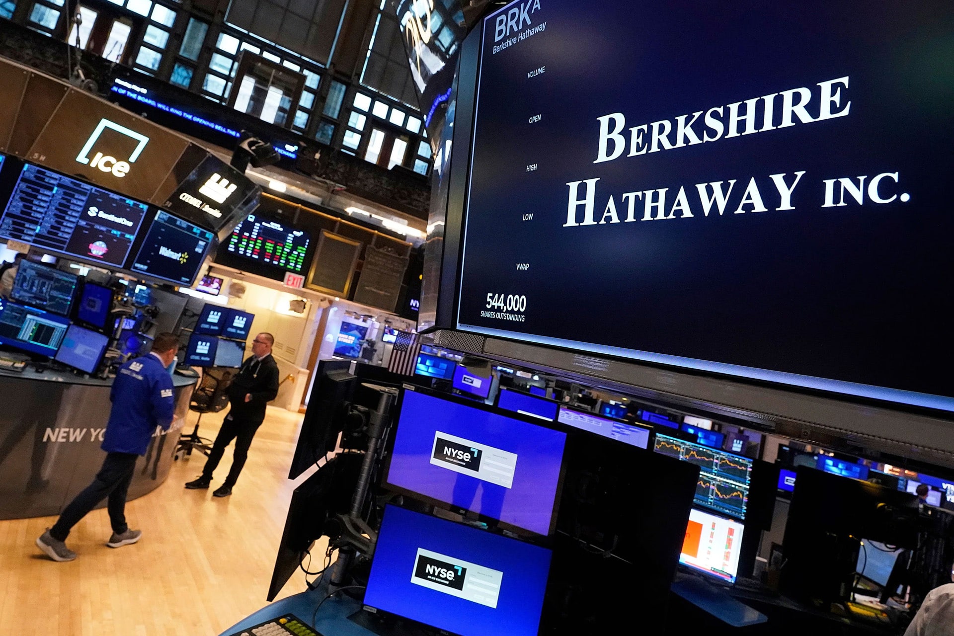 A stock exchange trading floor displays monitors, one prominently showing "Berkshire Hathaway Inc.," highlighting Warren Buffett's influence as traders walk nearby, surrounded by computer screens and financial data, including Apple stocks.