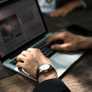 A person wearing a wristwatch is typing on a laptop at a wooden table, likely planning a business start. The laptop screen shows text, graphs, and a coffee cup image. Another person’s hands are partially visible in the background.