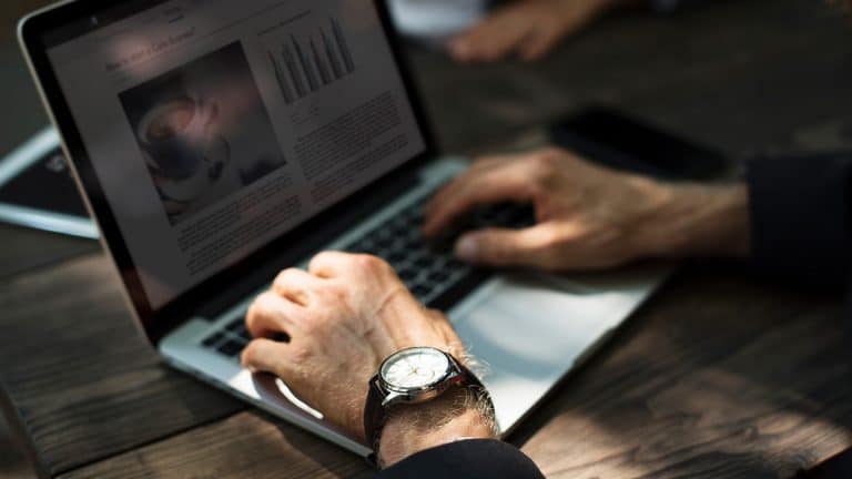 A person wearing a wristwatch is typing on a laptop at a wooden table, likely planning a business start. The laptop screen shows text, graphs, and a coffee cup image. Another person’s hands are partially visible in the background.