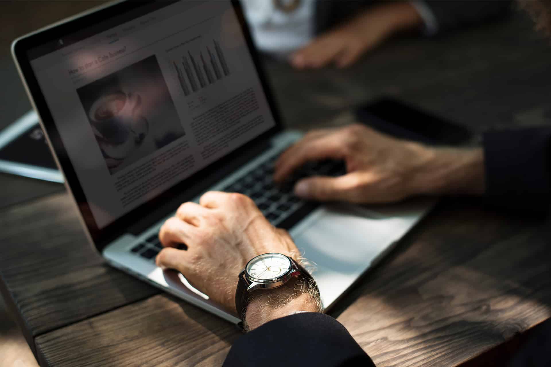 A person wearing a wristwatch is typing on a laptop at a wooden table, likely planning a business start. The laptop screen shows text, graphs, and a coffee cup image. Another person’s hands are partially visible in the background.