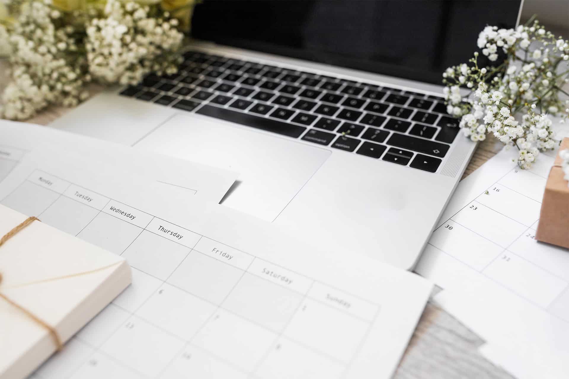 A laptop on a desk surrounded by paper calendars, a stack of envelopes tied with string, and white flowers creates an event planner's workspace that blends organization with decorative elements.
