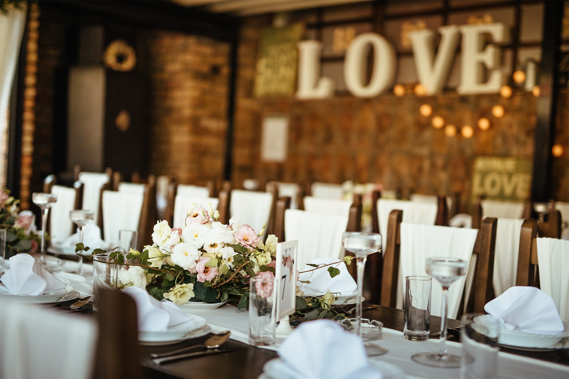 A long banquet table is elegantly set by an event planner with white napkins, glassware, and floral centerpieces. Large white letters spelling "LOVE" and warm string lights in the background create a romantic atmosphere.