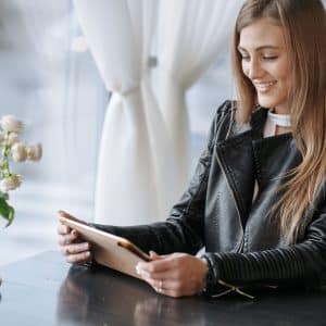 A woman with long hair, an event planner in a black leather jacket, sits at a table by a window, smiling while looking at a tablet. A vase with flowers is on the table beside her.