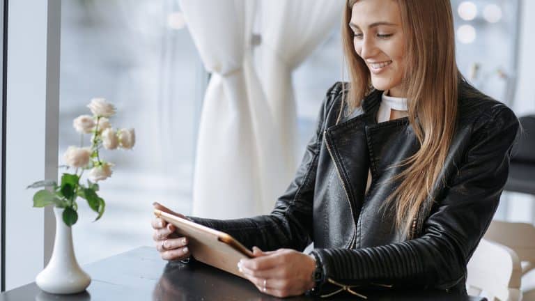 A woman with long hair, an event planner in a black leather jacket, sits at a table by a window, smiling while looking at a tablet. A vase with flowers is on the table beside her.