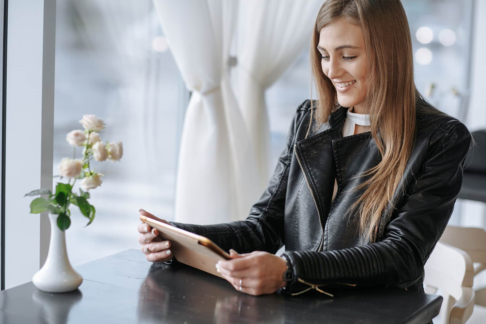 A woman with long hair, an event planner in a black leather jacket, sits at a table by a window, smiling while looking at a tablet. A vase with flowers is on the table beside her.