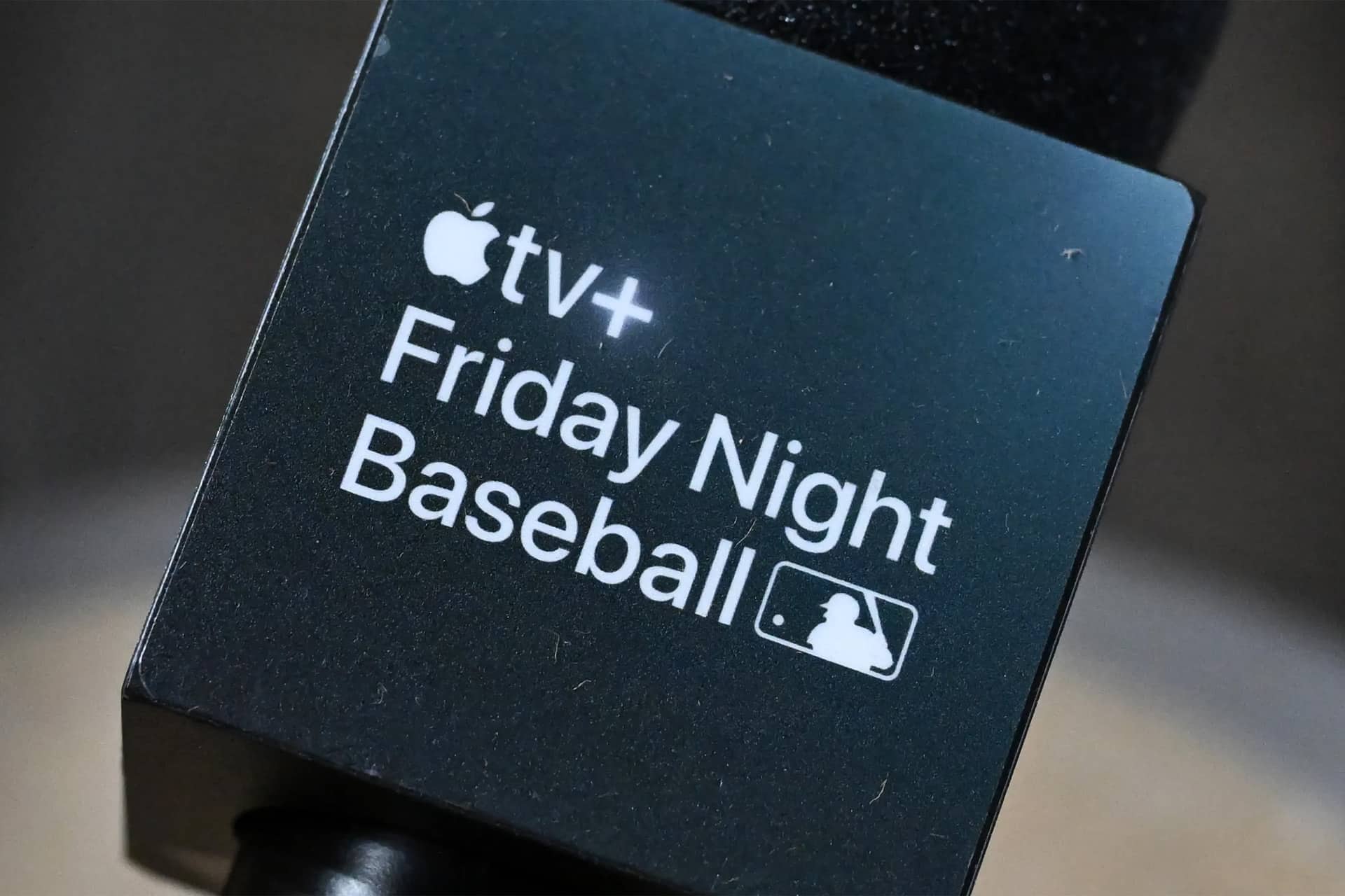 A close-up of a microphone with a black square label featuring the Apple TV+ logo and the text "Friday Night Baseball," positioned next to the Major League Baseball (MLB) logo.