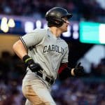A baseball player in a gray "Carolina" uniform and black helmet runs during a Friday Night Baseball game, with a blurred crowd and stadium lights in the background.