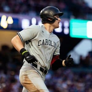 A baseball player in a gray "Carolina" uniform and black helmet runs during a Friday Night Baseball game, with a blurred crowd and stadium lights in the background.