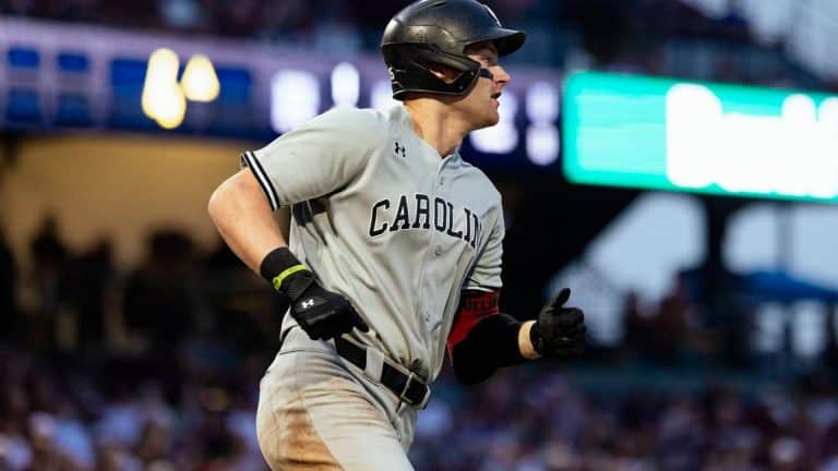 A baseball player in a gray "Carolina" uniform and black helmet runs during a Friday Night Baseball game, with a blurred crowd and stadium lights in the background.