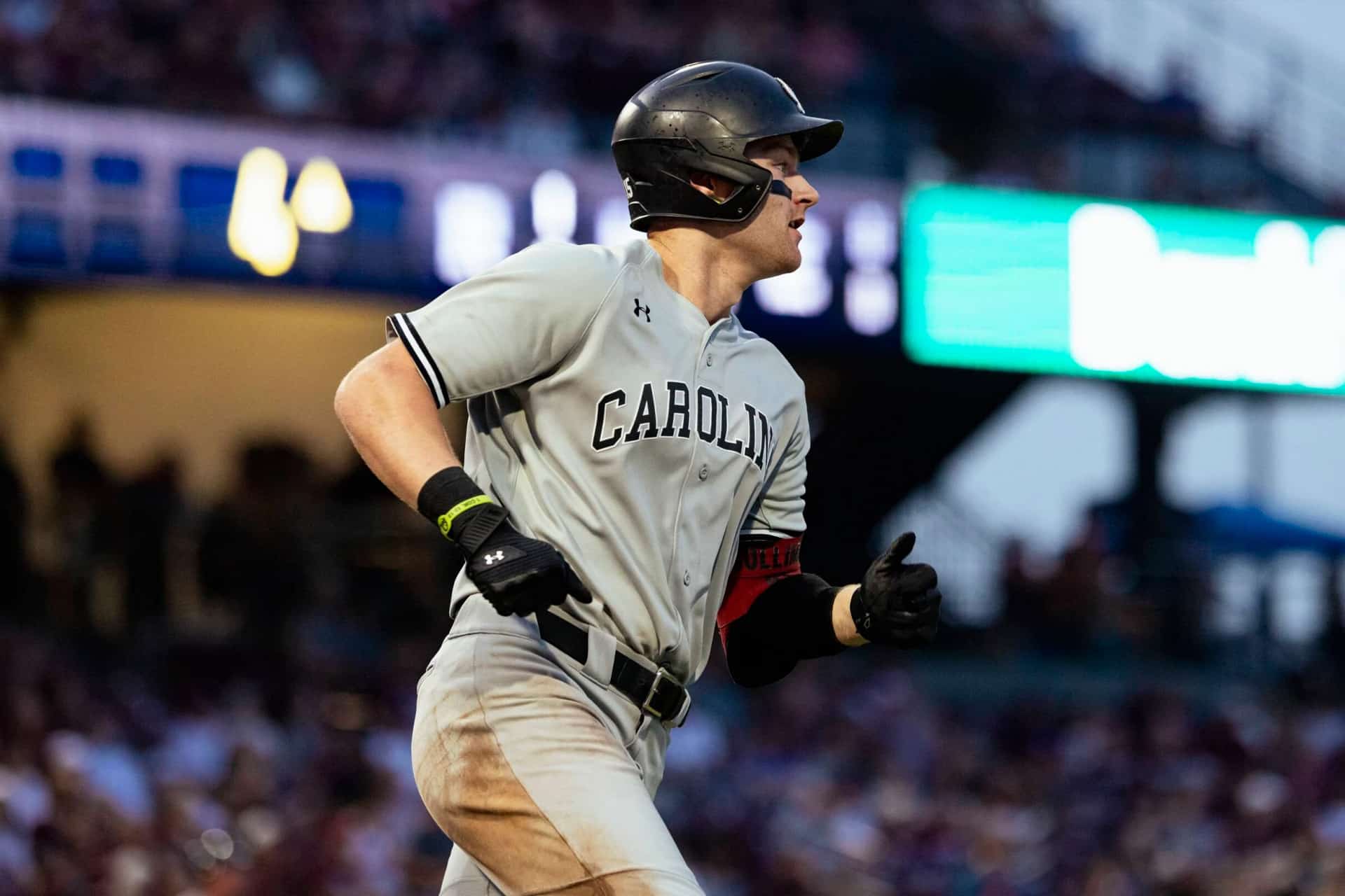 A baseball player in a gray "Carolina" uniform and black helmet runs during a Friday Night Baseball game, with a blurred crowd and stadium lights in the background.