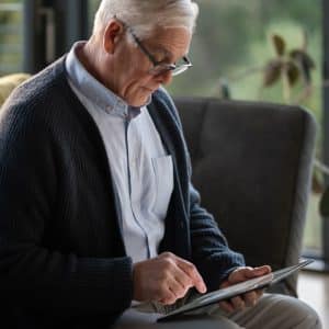An older man with gray hair and glasses sits on a sofa, using an Apple tablet. He wears a blue cardigan over a light shirt. There’s a yellow blanket beside him and a large window with greenery in the background.