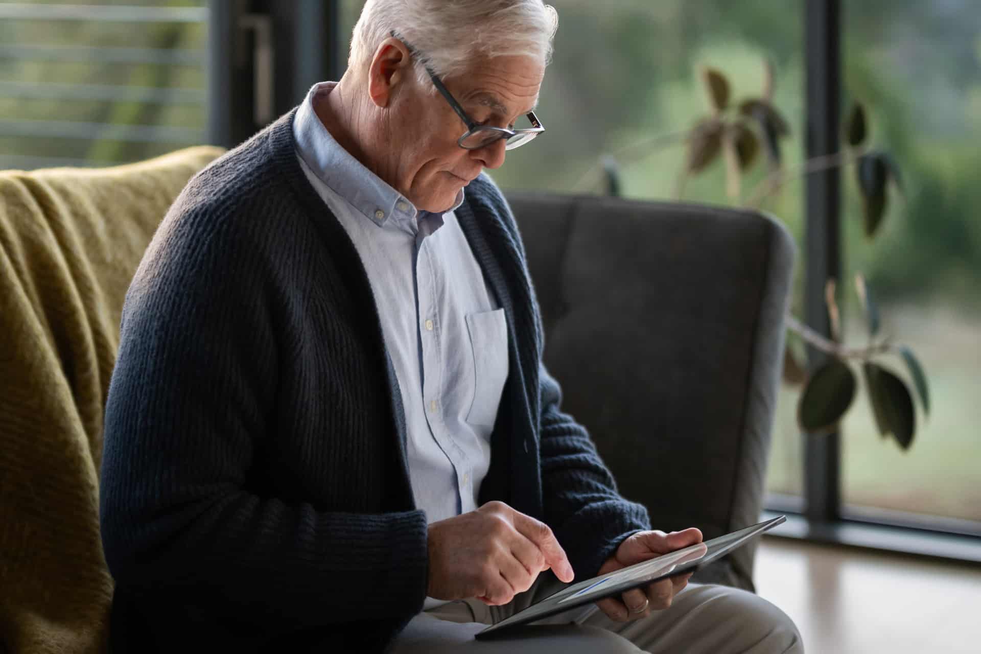 An older man with gray hair and glasses sits on a sofa, using an Apple tablet. He wears a blue cardigan over a light shirt. There’s a yellow blanket beside him and a large window with greenery in the background.