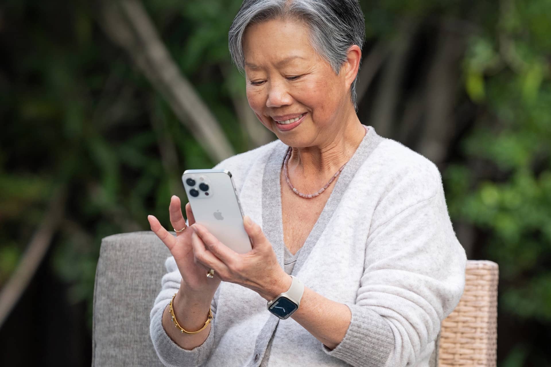 Apple Classroom - An older woman with gray hair smiles while using an Apple smartphone outdoors. She is wearing a light cardigan, a smartwatch, a bracelet, and a necklace, sitting on a wicker chair with greenery in the background.