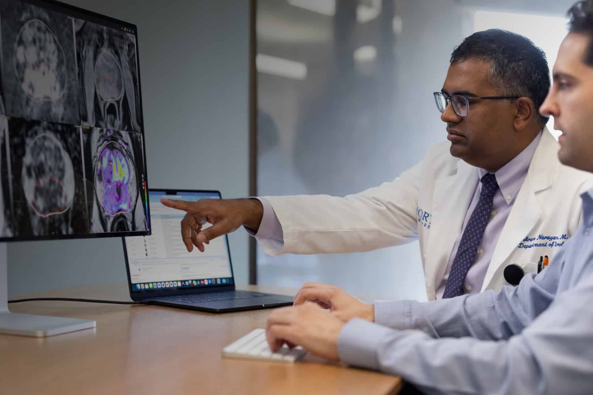Two doctors review DICOM medical imaging scans on a computer monitor. One, in a white lab coat, points at the screen while the other types. A Studio Display XDR laptop is open in the background on the table.