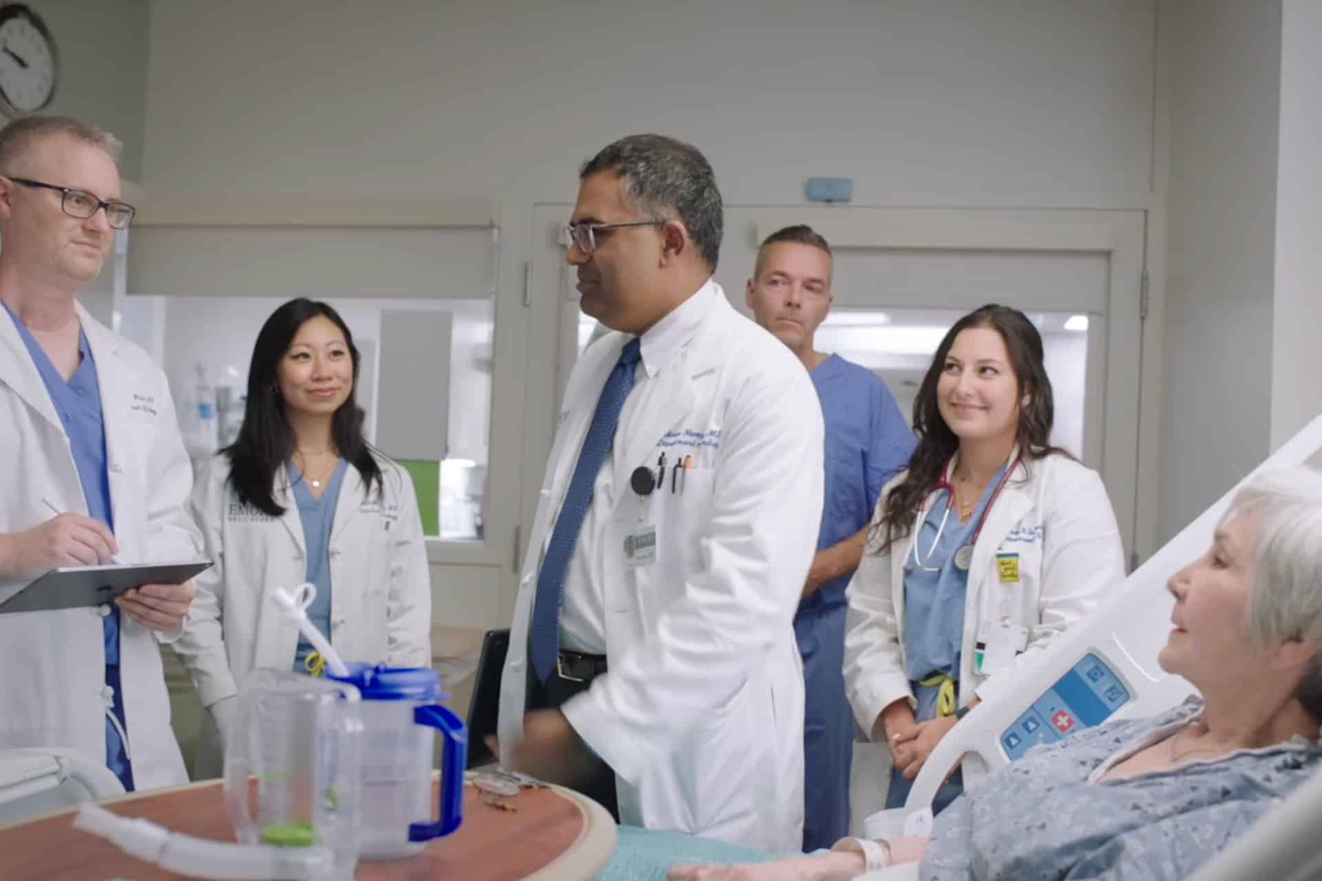 A group of doctors and nurses stand around a hospital bed, attentively listening to an elderly patient lying down. Medical equipment, a water pitcher, and an iPad monitoring the patient's vitals are visible in the foreground.