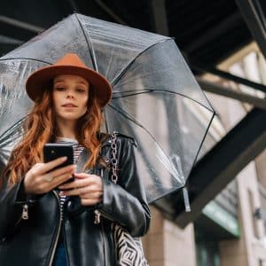 A young woman with long red hair wearing a brown hat and black leather jacket holds her smartphone, checking iPhone weather alerts as she stands under a clear umbrella on a city street.