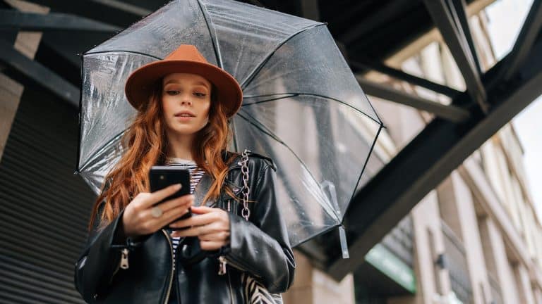A young woman with long red hair wearing a brown hat and black leather jacket holds her smartphone, checking iPhone weather alerts as she stands under a clear umbrella on a city street.