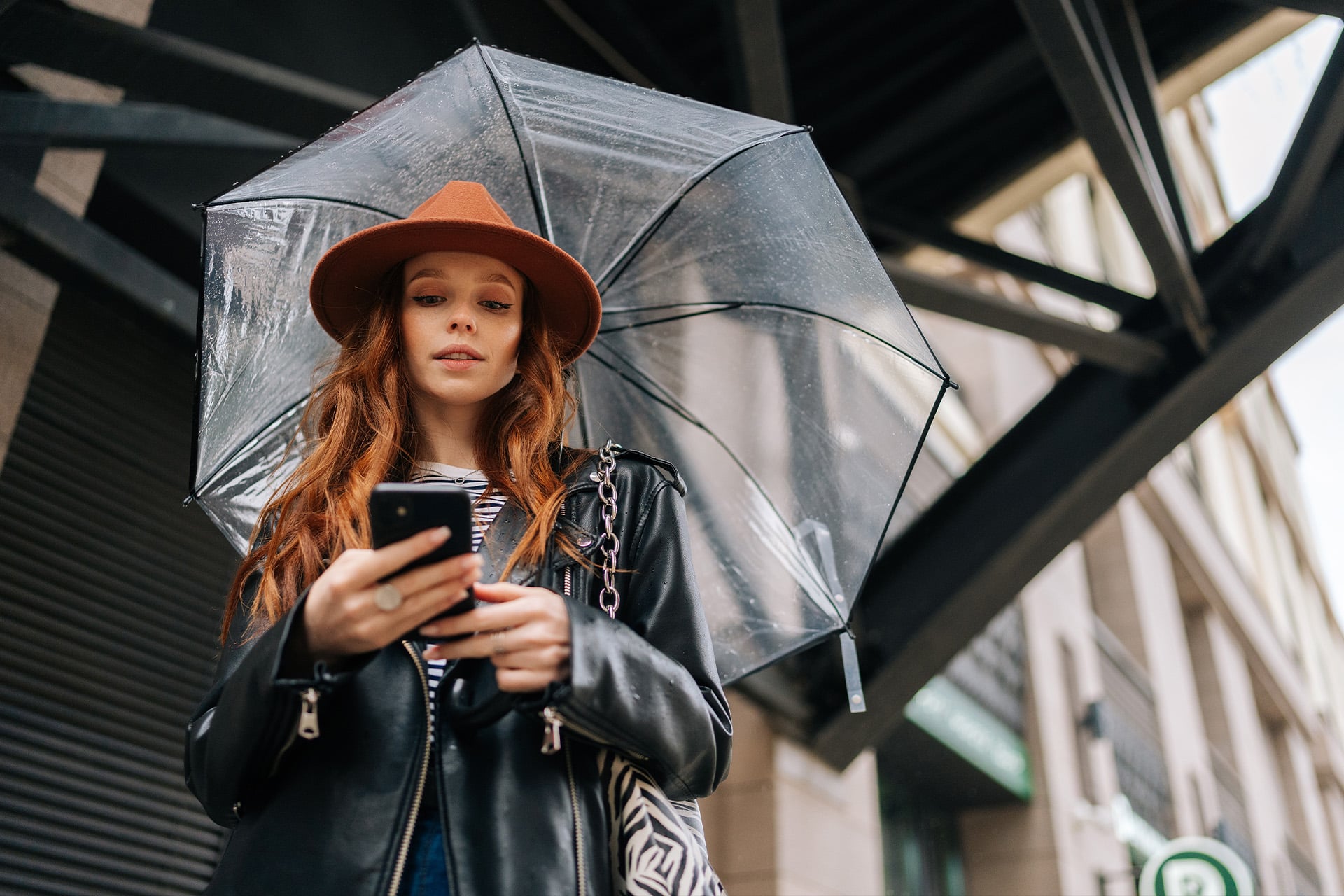 A young woman with long red hair wearing a brown hat and black leather jacket holds her smartphone, checking iPhone weather alerts as she stands under a clear umbrella on a city street.