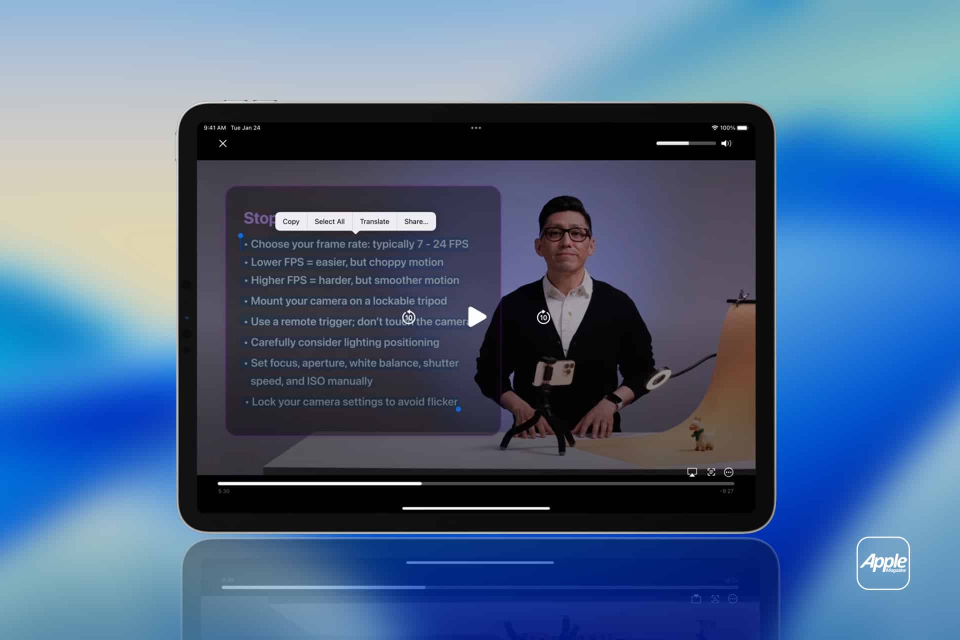 iPhone Live Text - A tablet displays a Life Text Video tutorial with a man standing next to a camera setup. A list of filming tips is visible on the screen. The blue gradient background features an Apple logo in the bottom right corner.