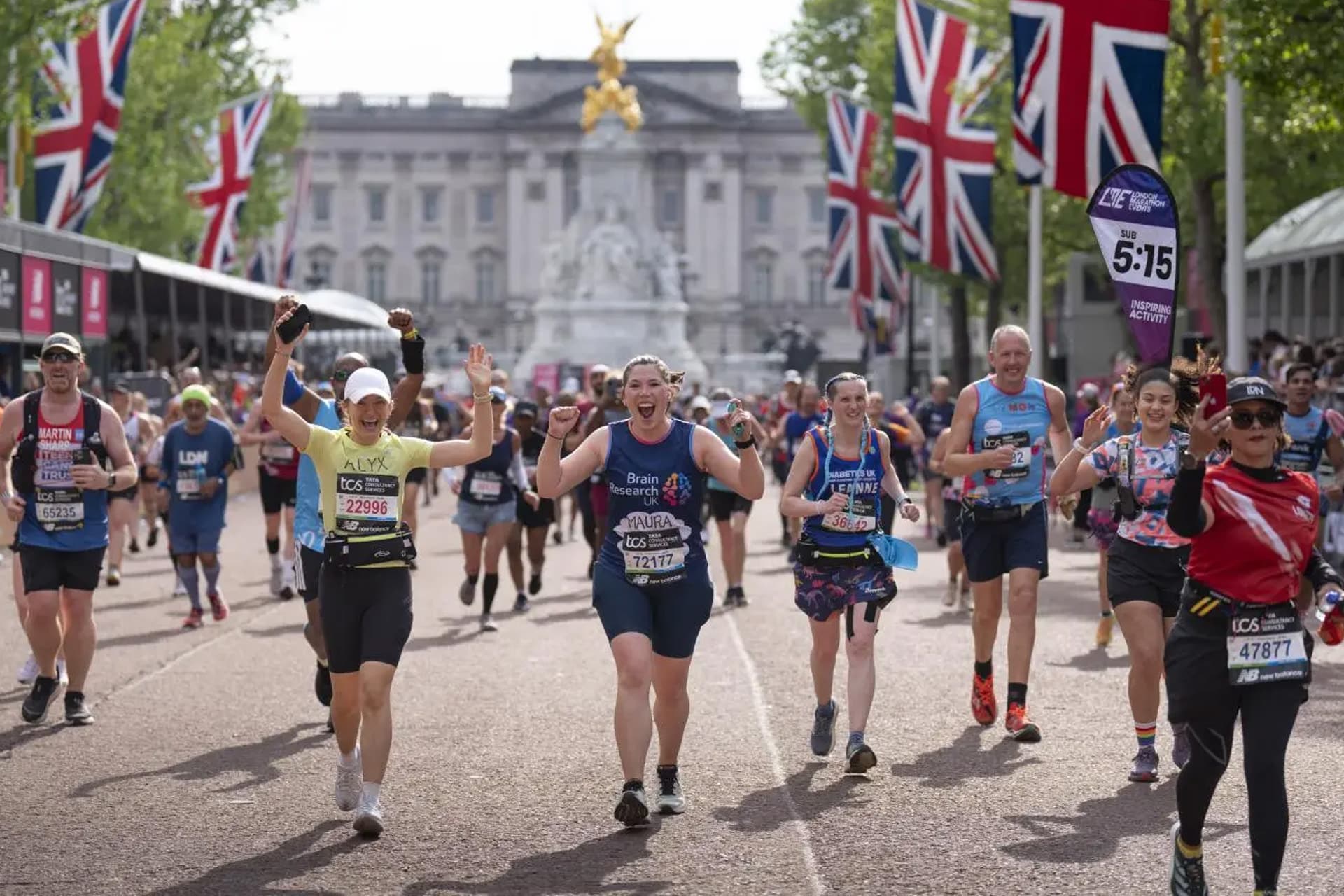 Runners celebrate as they approach the finish line of the London Marathon in front of Buckingham Palace, surrounded by Union Jack flags and cheering spectators on a sunny day.