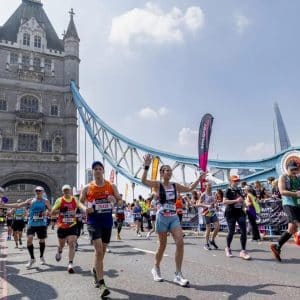 Runners participate in the London Marathon, crossing Tower Bridge on a sunny day, with crowds cheering on both sides and colorful banners lining the route.