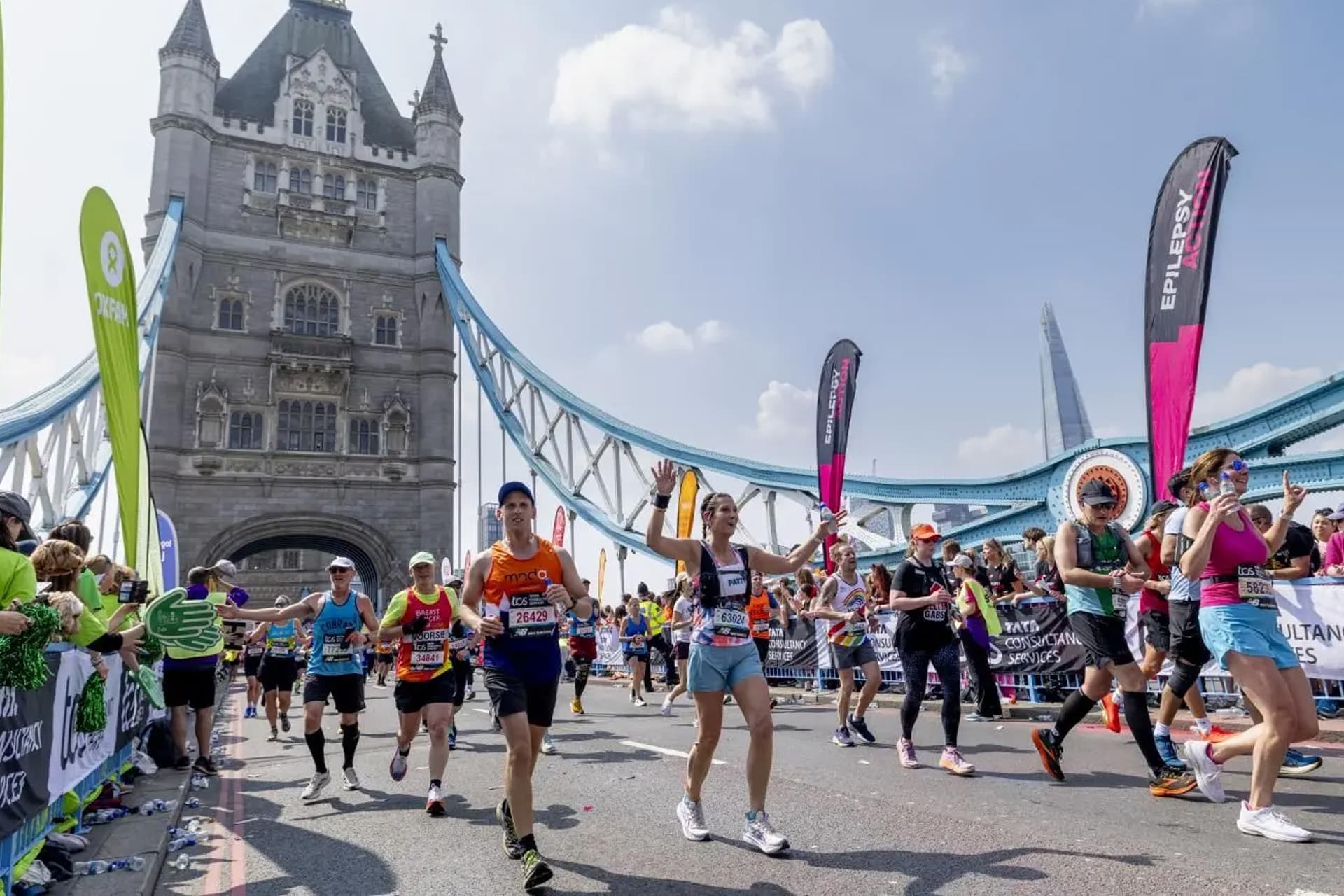 Runners participate in the London Marathon, crossing Tower Bridge on a sunny day, with crowds cheering on both sides and colorful banners lining the route.