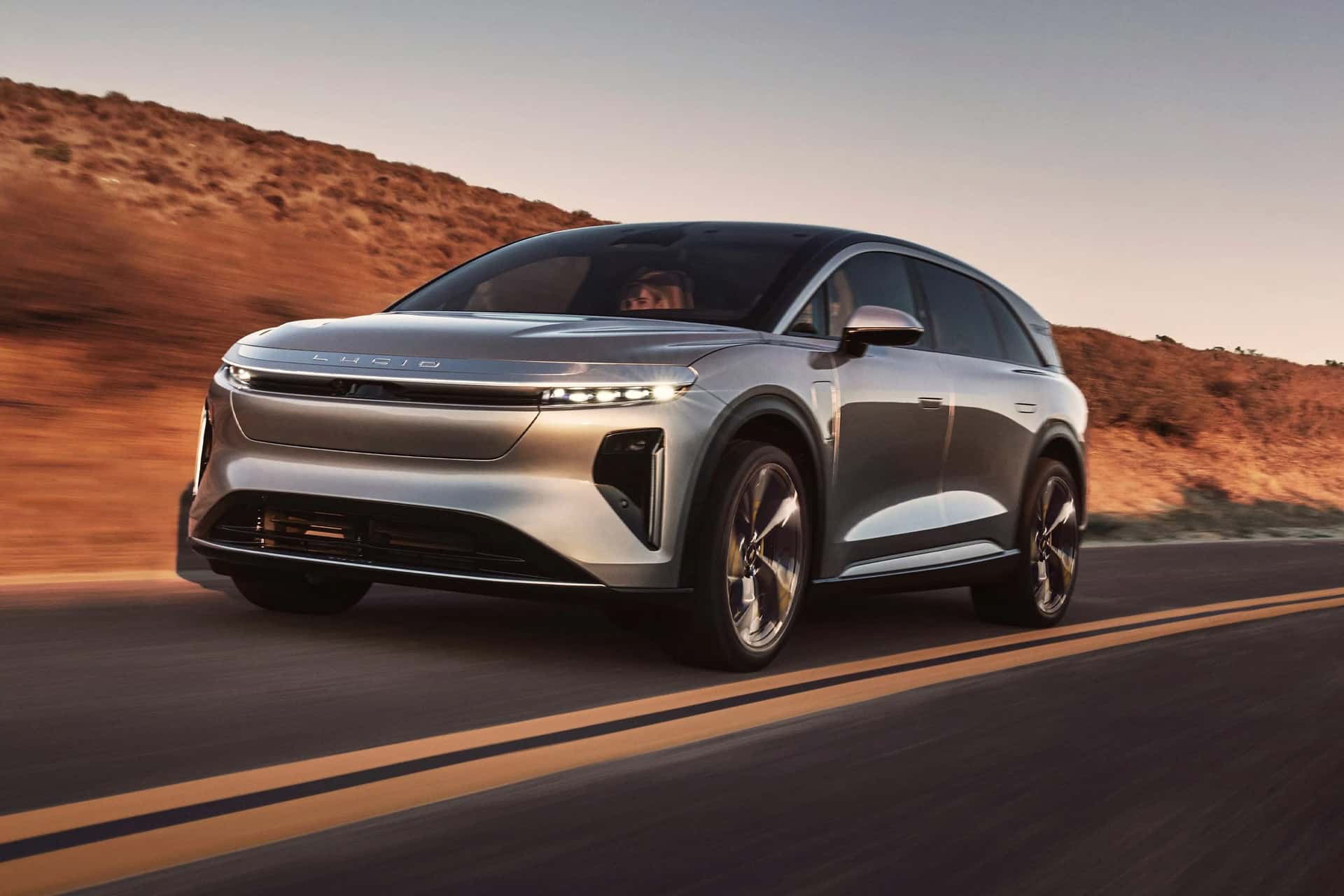 A silver Lucid Gravity SUV drives on a deserted road with hills and dry grass in the background during sunset, seamlessly integrating CarPlay for a connected journey.