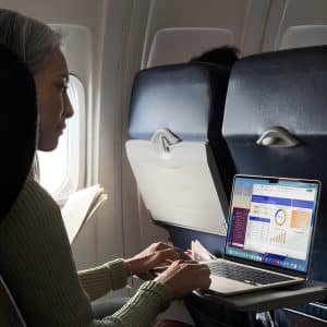 A woman sits on an airplane using a laptop on the tray table, viewing charts and graphs on the screen. She holds a document in her left hand and looks at the laptop, with sunlight coming through the window beside her.