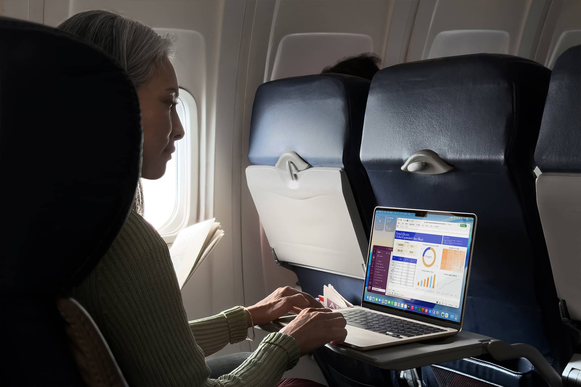 A woman sits on an airplane using a laptop on the tray table, viewing charts and graphs on the screen. She holds a document in her left hand and looks at the laptop, with sunlight coming through the window beside her.