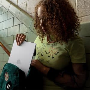 A young woman with curly hair stands on a stairway, sliding a silver laptop into a green backpack. She wears a light green T-shirt with cartoon characters and text, and is next to a tiled wall and stairs.