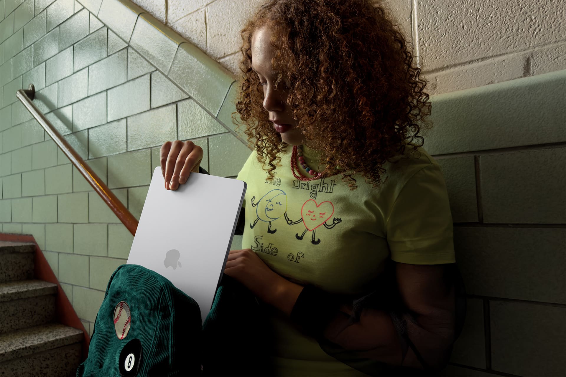 Student Research Workflow - A young woman with curly hair stands on a stairway, sliding a silver laptop into a green backpack. She wears a light green T-shirt with cartoon characters and text, and is next to a tiled wall and stairs.