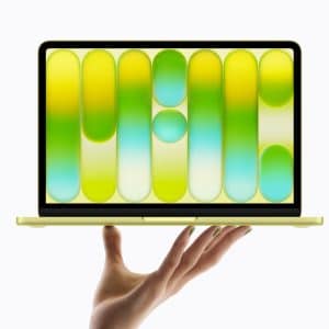 A hand holds up a thin, yellow laptop with a colorful abstract pattern of green, yellow, and blue shapes displayed on its screen against a white background.