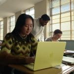 A student works on a yellow laptop at a desk in a sunlit classroom, while other students and a teacher interact in the background. Notebooks and pens are on the desk.