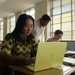 A student works on a yellow laptop at a desk in a sunlit classroom, while other students and a teacher interact in the background. Notebooks and pens are on the desk.
