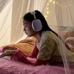 A girl wearing large white headphones lies on a fuzzy pink bedspread, using a laptop. She is under a sheer canopy, with soft pillows and string lights in the cozy background.