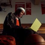 A person in sportswear sits on gym bleachers with a yellow laptop, a basketball, and an orange backpack nearby. Red championship banners hang on the wall in the background.